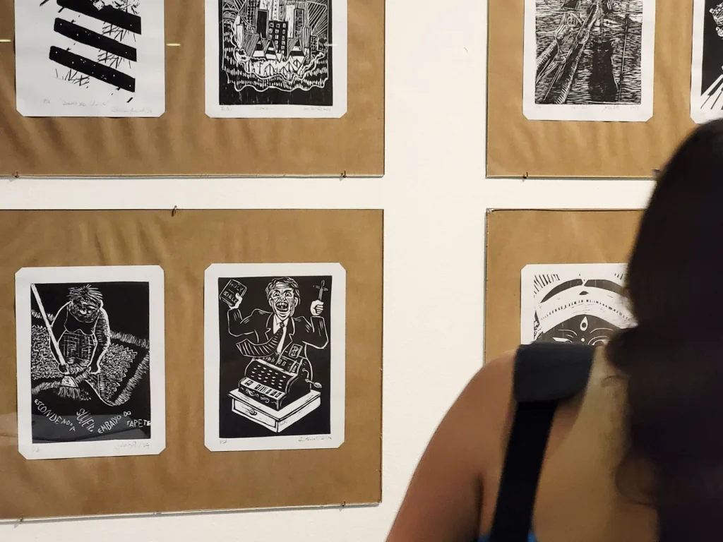 Costas de uma visitante da exposição em primeiro plano que olha paras as obras. Embaixo, à esquerda, se vê gravura de Antonio C. Silva mostrando um homem com uma cruz e uma bíblia saindo de uma caixa registradora. A de Julia Iguti está ao lado