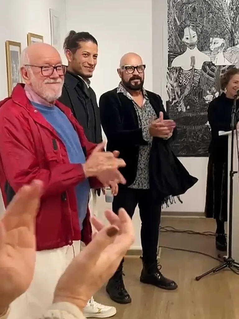 Artistas expositores em solenidade de abertura batendo palmas, próximo a um dos cantos da galeria, com obras ao fundo. Antonio em primeiro plano, com outros dois artistas homens. Ao fundo, a apresentadora fala em um púlpito